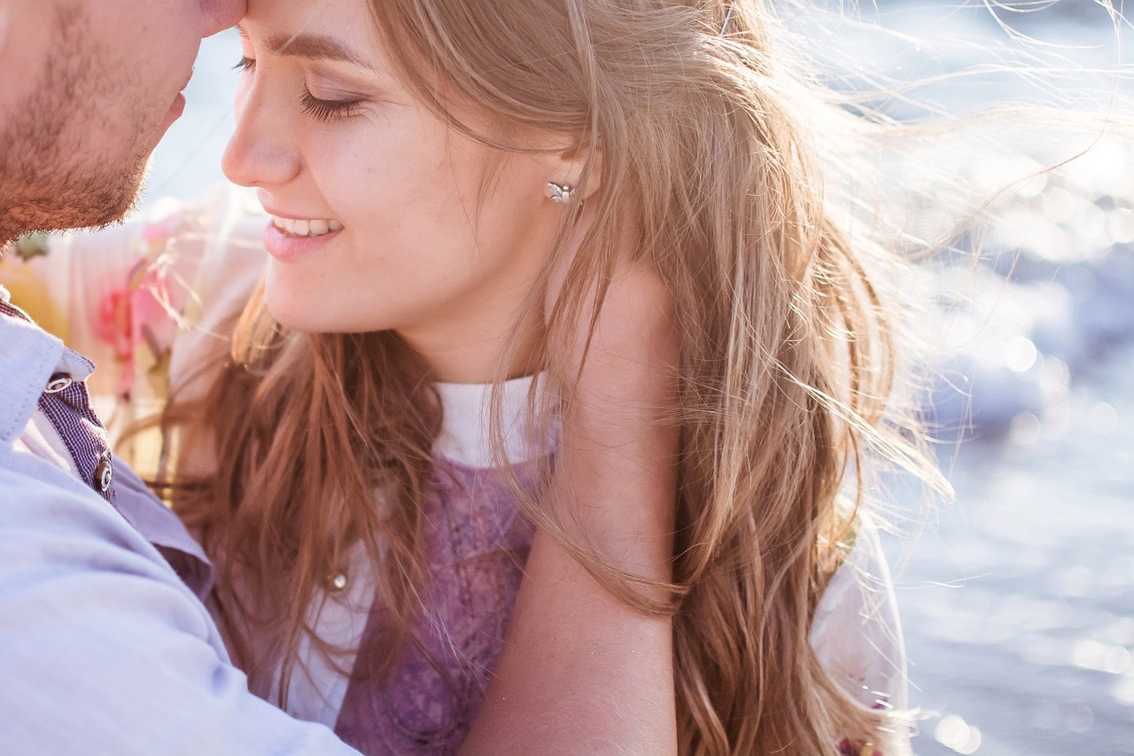 Man kissing a smiling woman's forehead beside sunlit water.